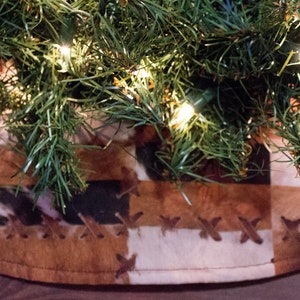 May include: A circular Christmas tree skirt featuring a patchwork design of brown, white, and black faux cowhide panels. The fabric sections are joined together with visible brown cross-stitch detailing along the seams, positioned under a pine tree.