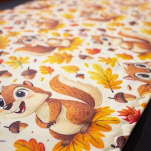 May include: A cream-colored table runner featuring a repeating pattern of cartoon chipmunks, autumn leaves, and acorns. The chipmunks are brown and tan with white bellies. The leaves are yellow, orange, and red. A decorative item for the fall season.