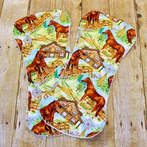 May include: Two cloth pads with a repeating pattern of brown horses, wooden barns, and windmills on a light blue and green background. The pads have a white trim and are arranged on a wooden surface.