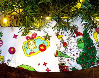 May include: White Christmas tree skirt featuring a colorful cartoon design with the Grinch, Cindy Lou Who, and other characters. The skirt has a scalloped edge and is decorated with stars and ornaments. The Grinch is shown with a Christmas tree.