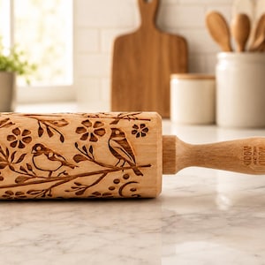 May include: A light-coloured wooden rolling pin featuring an intricate laser-engraved pattern of birds perched on floral branches. The handle is stamped with the text WOOD WOOD. The surface of the rolling pin is smooth and cylindrical in shape.