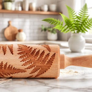 A wooden rolling pin with an engraved fern leaf pattern rests on a light marble countertop in a bright kitchen. In the background, a white vase with green fern leaves and light-colored kitchen furnishings create a cozy, natural atmosphere.
