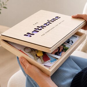 Könnte beinhalten: Eine hellfarbene Holz-Andenkenbox mit dem Namen "Katherine" auf dem Deckel ausgeschnitten. Die Box ist geöffnet und zeigt Fotos, Schlüssel und andere Gegenstände. Der Text "FOR EVERYTHING THAT MATTERS" befindet sich ebenfalls auf dem Deckel.