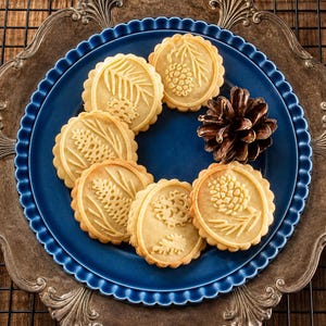 May include: A blue plate with a scalloped edge holds several round, light-coloured cookies with embossed botanical designs. A pine cone and a decorative silver tray are also present. A wooden spoon rests on the side.
