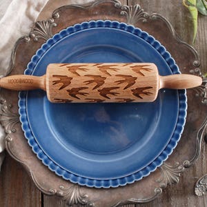 以下が含まれることがあります： 鳥が彫刻された木製麺棒。麺棒は銀色の縁取りのある青い皿の上に置かれています。皿は木製のテーブルの上に置かれています。