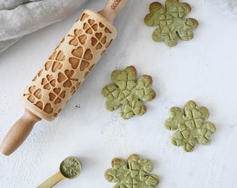 Pode incluir: Um rolo de massa de madeira com um design de trevo, uma pequena colher de pó verde e cinco biscoitos verdes em forma de trevo sobre uma superfície branca. O rolo de massa é gravado com as palavras "WOODGOOD".