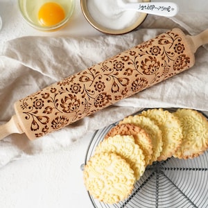 Wooden rolling pin with decorative design of polish folk traditional flowers. A well baked cookies presenting the effects of embossing are lying on a metal trivet  in front of it.