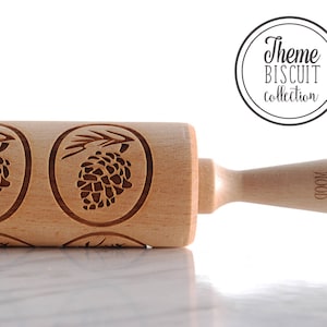 May include: A wooden rolling pin with a pine cone design. The rolling pin has a handle with the words "Mood Wood" engraved on it. The rolling pin is on a white surface.