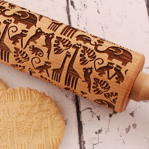 Wooden rolling pin laser engraved with design of tropical animals such as elephants, giraffs, parrots, monkeys, surrounded by palm and monstera leaves. Three big roundcookies are lying in front of it.