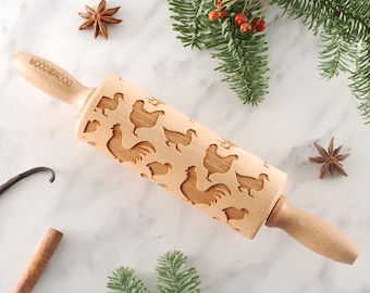 May include: A wooden rolling pin with a chicken design engraved on the surface. The rolling pin is on a white marble surface with a sprig of evergreen, cinnamon sticks, and star anise.