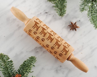 May include: A wooden rolling pin with a design of dogs and paw prints. The rolling pin is on a white marble surface with green pine branches and a star anise.