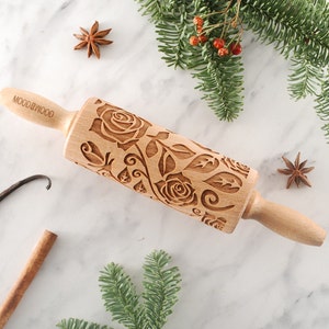 Pode incluir: Um rolo de massa de madeira com um design floral com rosas e folhas. O rolo é gravado com o texto "MOODBYWOOD".