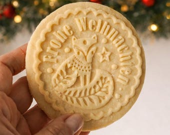 May include: A round, light-colored cookie with a scalloped edge, featuring an embossed design of a fox surrounded by decorative patterns and the German text Frohe Weihnachten, which translates to Merry Christmas in English.