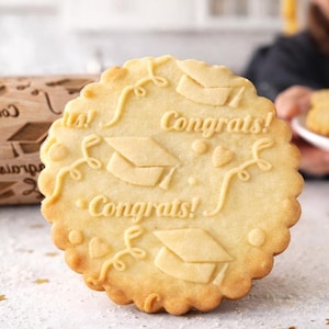 High-quality product photo showing a wooden engraved rolling pin with a detailed graduation-themed pattern, including caps, hearts, and “Congrats” text. A realistic magnifying glass highlights the exact same engraving, revealing deep, precise carving and natural wood grain texture. Surrounding baking elements like eggs, flour, and freshly baked embossed cookies create a warm, artisanal kitchen scene. Soft natural lighting and shallow depth of field emphasize craftsmanship and premium quality.