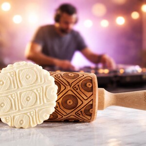 May include: A wooden rolling pin with a record player design is displayed next to a cookie cut in the same shape. The rolling pin has a light wood handle and the cookie is a light beige color.