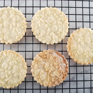 May include: Five round, white sugar cookies with a floral design are cooling on a black wire rack. The cookies are slightly browned on the edges.