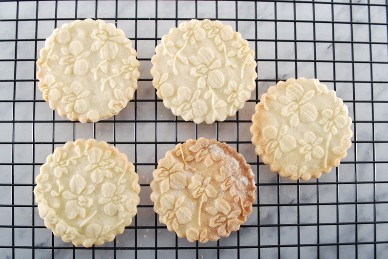 May include: Five round, white sugar cookies with a floral design are cooling on a black wire rack. The cookies are slightly browned on the edges.