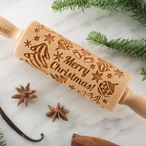 May include: A wooden rolling pin with a Christmas design. The rolling pin is engraved with the words "Merry Christmas!" and features images of Christmas trees, stars, bells, and ornaments. The rolling pin is on a white marble surface.