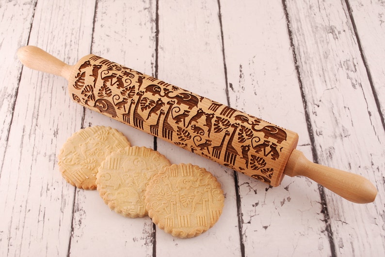 Wooden rolling pin laser engraved with design of tropical animals such as elephants, giraffs, parrots, monkeys, surrounded by palm and monstera leaves. Three big roundcookies are lying in front of it.