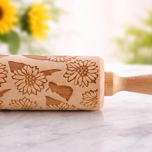 May include: A wooden rolling pin with a sunflower pattern carved into the surface. The rolling pin is light brown and has a handle on one end. The background is blurred, with yellow sunflowers visible.