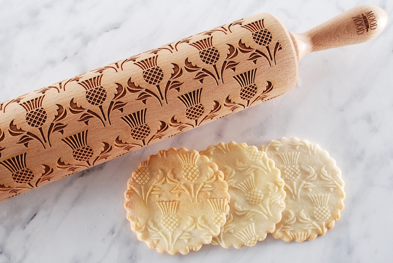 Wooden rolling pin with moodforwood logo on handle, laser engraved with design of scottish thistle repetitive design. A round well baked cookies with embossed design are lying in front of it.