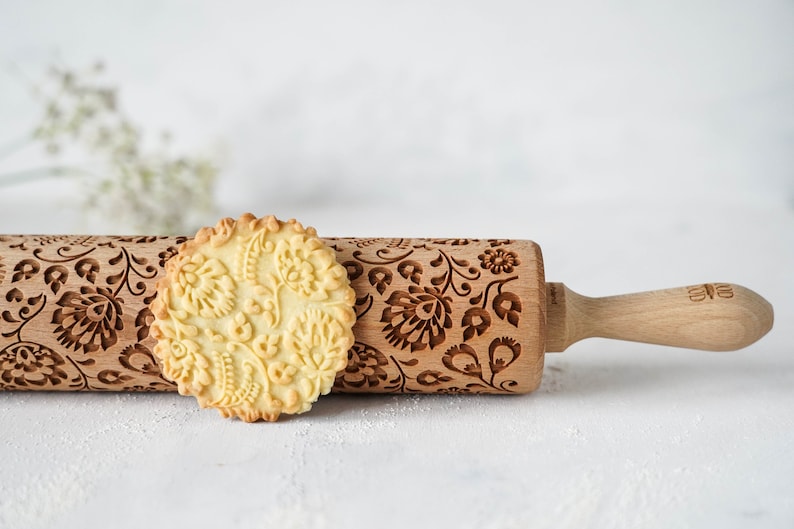 Wooden rolling pin with decorative design of polish folk traditional flowers. A well baked cookie presenting the effects of embossing is standing in front of it.