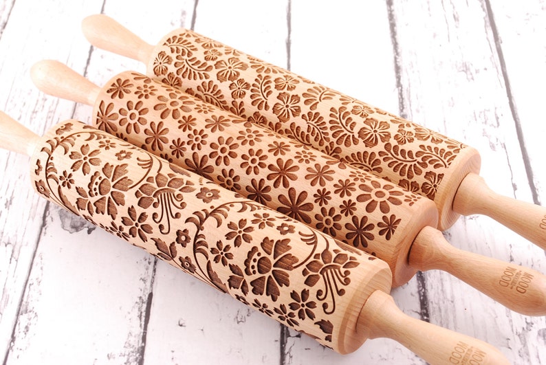 three wooden rolling pins laser engraved with 3 different floral patterns. They are on white board rustic background.