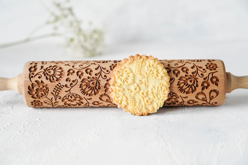 Wooden rolling pin with decorative design of polish folk traditional flowers. A well baked cookie presenting the effects of embossing is standing in front of it.