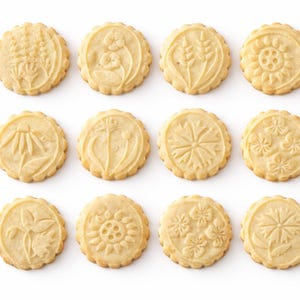 Minimalist product composition featuring nine embossed botanical cookies arranged in a neat grid on a bright white background. On the right, elegant serif headline reads “Real baking results,” followed by a refined benefits list with icons, highlighting texture, timeless design, uniqueness, gifting appeal, customization, and creative baking. Soft shadows, warm tones, and luxury-style typography create a premium, high-end visual feel.