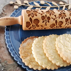 May include: A wooden rolling pin with a floral design, engraved with the words "MOOD & WOOD". The rolling pin is on a blue plate with four cookies that have been rolled with the pin, creating a floral pattern on the cookies.