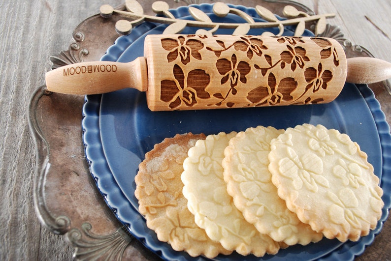 May include: A wooden rolling pin with a floral design, engraved with the words "MOOD & WOOD". The rolling pin is on a blue plate with four cookies that have been rolled with the pin, creating a floral pattern on the cookies.