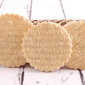 May include: Three round, light brown cookies with the text "Amelie's cookies" repeated on each cookie. The cookies are decorated with small heart shapes.