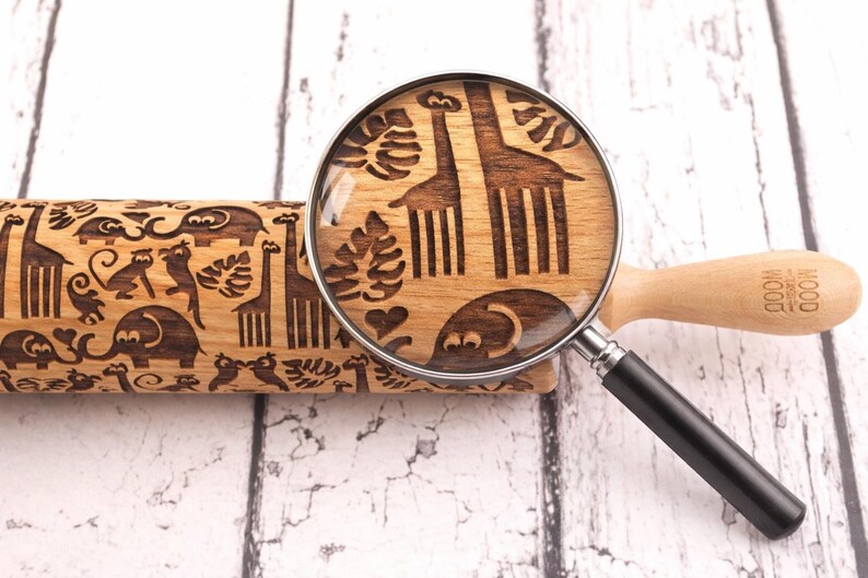 A close-up image of a wooden embossed rolling pin featuring a playful safari pattern with animals like giraffes, elephants, and monkeys. A magnifying glass highlights the intricate engraved details on the surface, emphasizing the precision and craftsmanship of the design. The rolling pin rests on a light, rustic wooden background, creating a clean and natural aesthetic that draws attention to the decorative pattern.