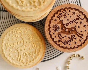 May include: Close-up of Christmas-themed cookies and a wooden stamp. The cookies are round with embossed designs of a reindeer, stars, and the words "Joyeux Noël". The wooden stamp features a reindeer and the words "Joyeux Noël" in a decorative font.