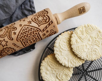 Pode incluir: Um rolo de madeira com desenhos de feriados gravados, incluindo um homem de gengibre e uma casa, ao lado de biscoitos em relevo em uma grelha. Os biscoitos apresentam padrões festivos semelhantes. O rolo é feito de madeira clara.