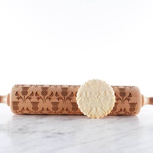 Wooden rolling pin with moodforwood logo on handle, laser engraved with design of scottish thistle repetitive design. A round well baked cookies with embossed design are lying in front of it.