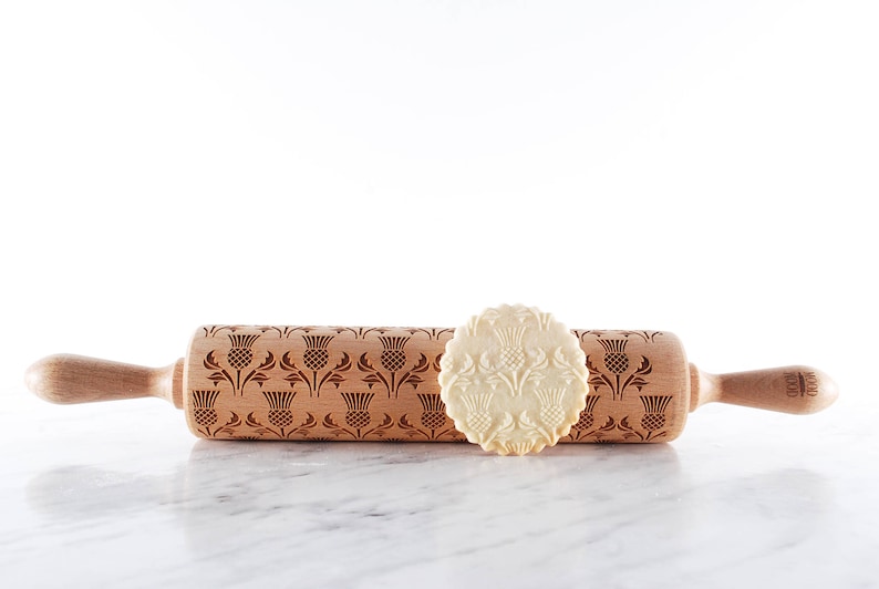 Wooden rolling pin with moodforwood logo on handle, laser engraved with design of scottish thistle repetitive design. A round well baked cookies with embossed design are lying in front of it.