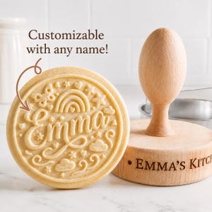 May include: A round wooden cookie stamp with a light wood finish, featuring an embossed design of a rainbow, stars, clouds, and a butterfly surrounding the name Emma in script. The text Customizable with any name! is visible above the stamp.