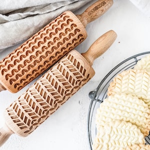 May include: Two wooden rolling pins with different engraved patterns. One has a wavy pattern and the other has a chevron pattern. The rolling pins are on a white surface. There are cookies on a wire rack in the background.