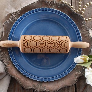 Wooden small rolling pin laser engraved with design of honeycomb, bees, hearts. For embossing cookies. It is lying on a blue plate and metal dish.