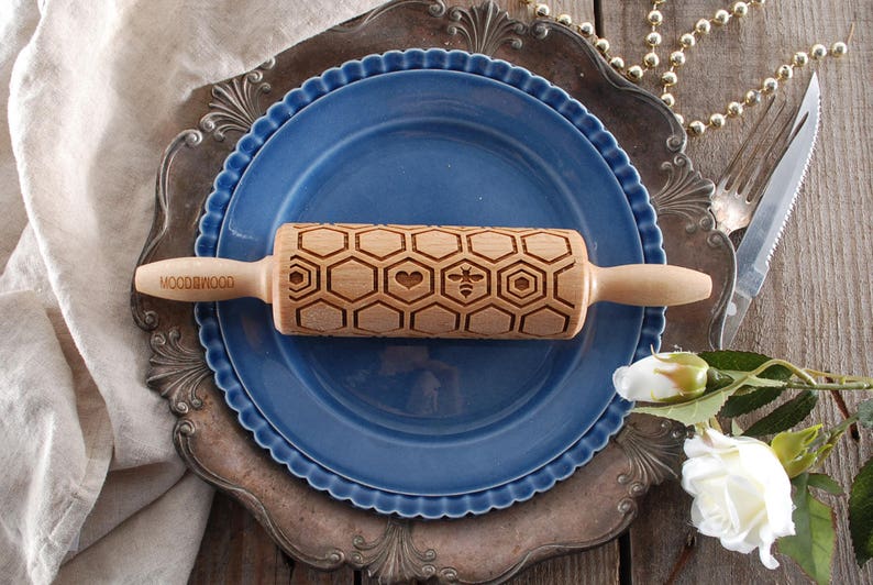 Wooden small rolling pin laser engraved with design of honeycomb, bees, hearts. For embossing cookies. It is lying on a blue plate and metal dish.