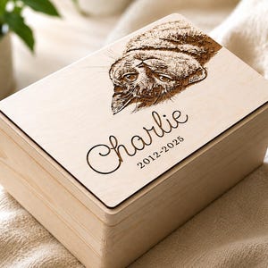 Op de afbeelding: Een lichtgekleurde houten kist met een gegraveerd kattenportret op het deksel. De naam "Charlie" en de jaren "2012-2025" zijn ook gegraveerd. De kist staat op een crèmekleurige stof.