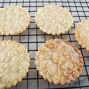 May include: Five round, white sugar cookies with an embossed floral design are arranged on a black wire rack. The cookies are slightly browned on the edges.