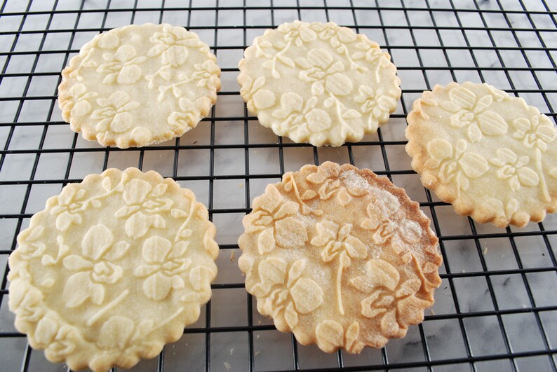 May include: Five round, white sugar cookies with an embossed floral design are arranged on a black wire rack. The cookies are slightly browned on the edges.