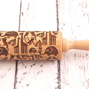 Wooden rolling pin laser engraved with design of tropical animals such as elephants, giraffs, parrots, monkeys, surrounded by palm and monstera leaves.
