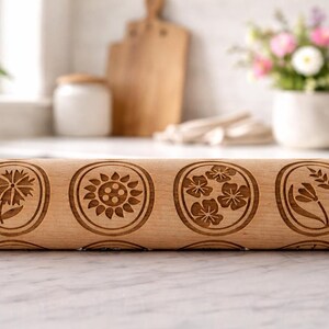 Bright, airy kitchen scene featuring a wooden embossed rolling pin with floral patterns placed on a marble countertop. The background is softly blurred, showing a white ceramic pot filled with fresh flowers, a wooden cutting board, and minimal kitchen elements. Natural light enhances the warm tones of the wood and highlights the intricate engraved details, creating a calm, elegant, and inviting atmosphere.
