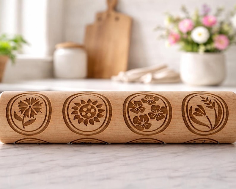 Bright, airy kitchen scene featuring a wooden embossed rolling pin with floral patterns placed on a marble countertop. The background is softly blurred, showing a white ceramic pot filled with fresh flowers, a wooden cutting board, and minimal kitchen elements. Natural light enhances the warm tones of the wood and highlights the intricate engraved details, creating a calm, elegant, and inviting atmosphere.