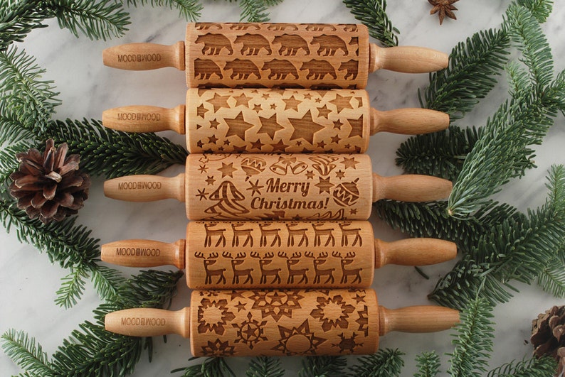 Five small wooden rolling pins with laser engraed designs on them: snowflakes, reindeer, merry christmas text, stars and polar bears. Among christmas tree branches.