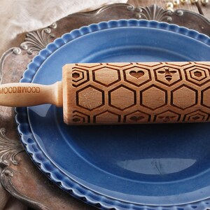 Wooden small rolling pin laser engraved with design of honeycomb, bees, hearts. For embossing cookies. It is lying on a blueplate and metal dish.