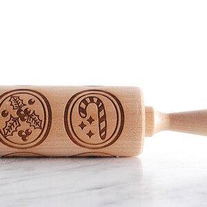 Wooden embossed rolling pin with festive holiday patterns placed on a light marble surface, with three freshly baked cookies in front showing detailed imprints such as holly and a snowflake. In the background, softly blurred Christmas lights and decorated trees create a warm golden bokeh effect. The scene highlights the natural wood texture, precise engraving, and cozy seasonal atmosphere, combining product detail with a festive, high-end lifestyle setting.